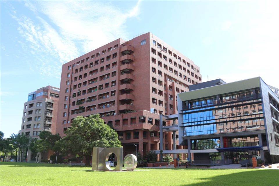 晴朗藍天下的成大校園，一棟紅磚高樓旁是玻璃帷幕的現代建築，前方是寬廣的草坪與大型金屬字母雕塑。