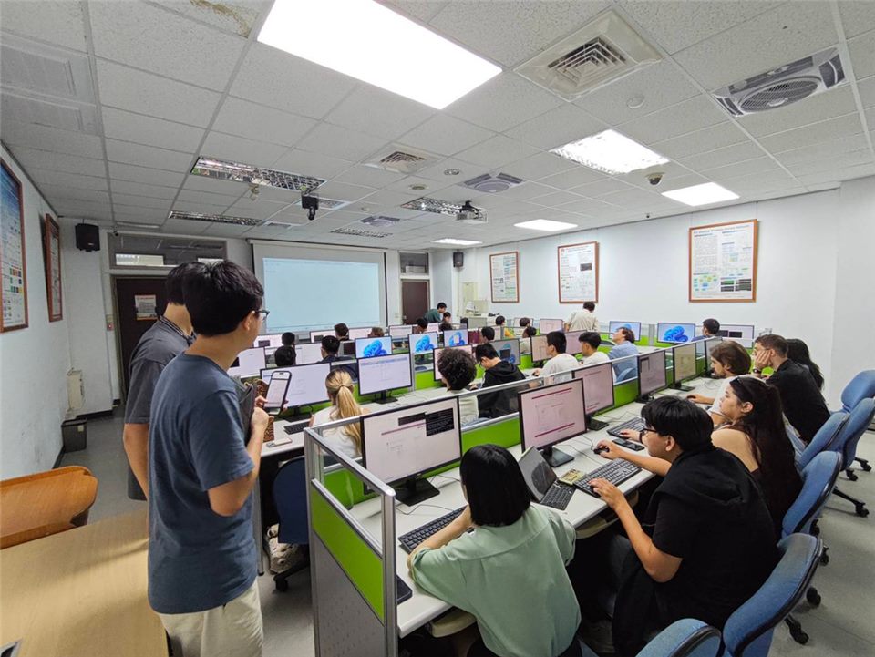 電腦教室裡，學員坐在一排排以綠色隔板區隔的電腦前操作，一位學員站在走道旁看著手機。