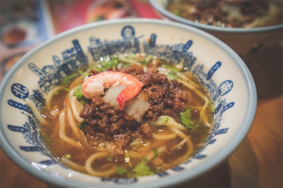 台南擔仔麵：藍白花紋的碗裡是黃色油麵，上面鋪著肉燥、一隻蝦子與香菜。