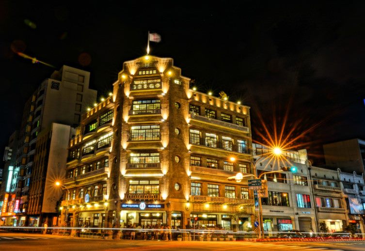 夜晚燈火通明的多層樓老建築，外牆亮起一圈圈暖黃色燈光，屋頂立著旗桿，前方街道有車燈劃出的光軌。